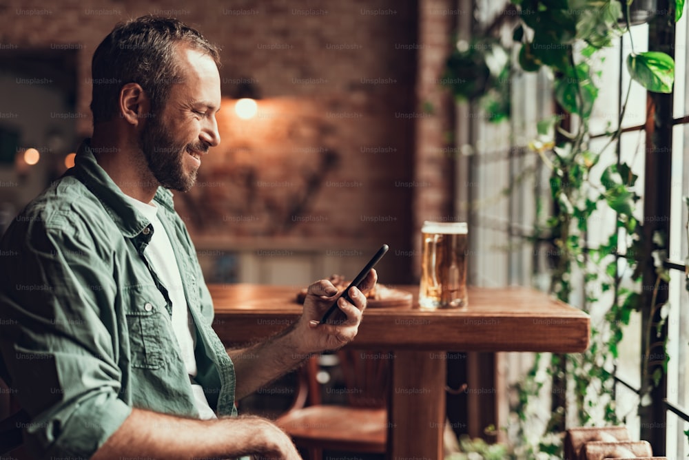 Retrato de vista lateral de un joven y guapo caballero revisando mensajes en el teléfono inteligente mientras está sentado en una silla. Está mirando la pantalla del teléfono y sonriendo