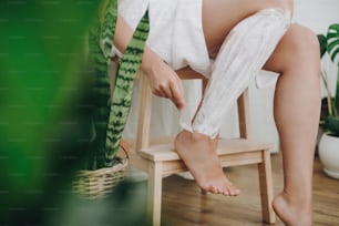 Young woman in white towel applying shaving cream on her legs in home bathroom with green plants. Skin care and wellness concept. Hand holding plastic razor on skin with cream