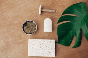 Zero waste flat lay. Natural soap, solid shampoo in metal tin, reusable razor, crystal eco deodorant, sponge on wooden background with green monstera leaf. Plastic free beauty essentials