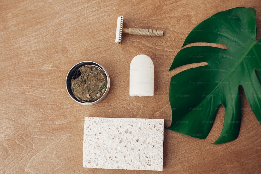 Zero waste flat lay. Natural soap, solid shampoo in metal tin, reusable razor, crystal eco deodorant, sponge on wooden background with green monstera leaf. Plastic free beauty essentials