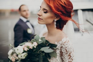 sensual stylish bride holding modern bouquet and relaxing with windy hair and groom looking at her near retro car. luxury wedding couple newlyweds posing. space for text. emotional moment