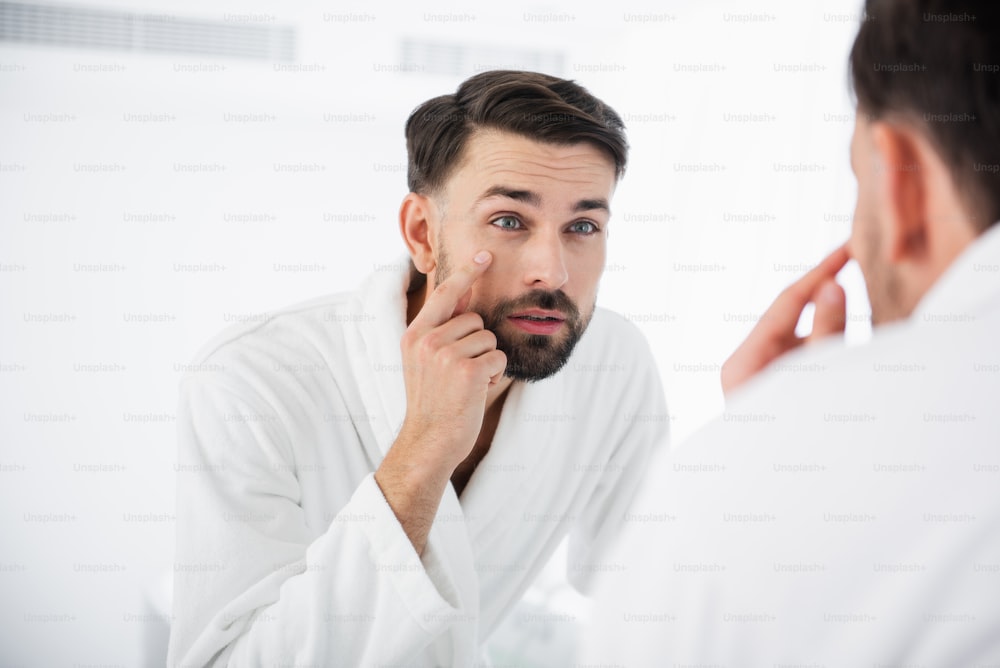 Calm bearded middle aged man thoughtfully leaning to the mirror while touching the cheek with one finger