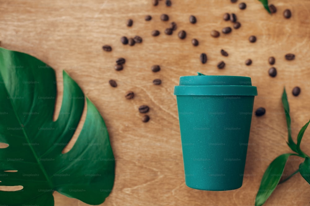 Stilvolle wiederverwendbare Öko-Kaffeetasse auf Holzhintergrund mit gerösteten Kaffeebohnen und grünem Monsterablatt. Verbot von Einwegkunststoff, Zero-Waste-Konzept, flache Verlegung. Nachhaltiger Lebensstil.