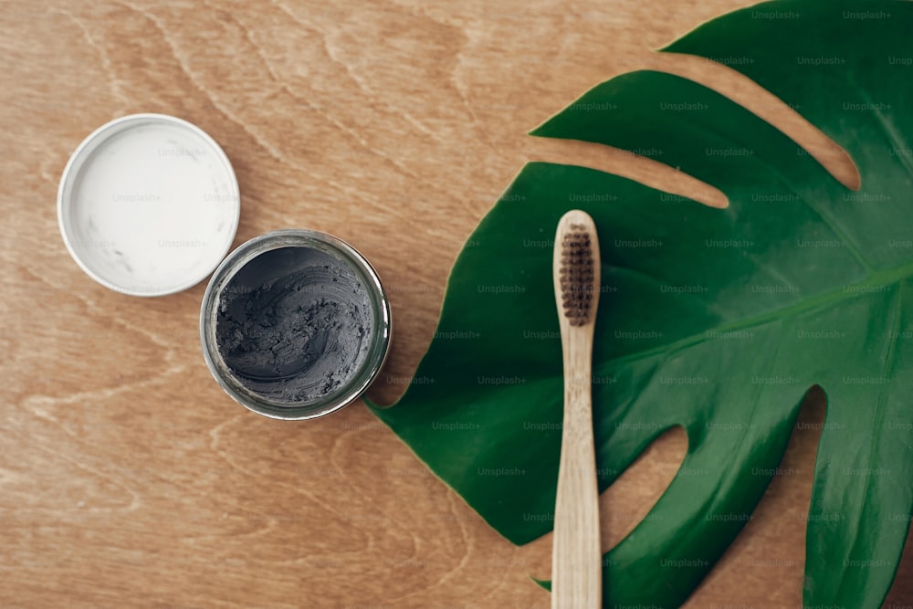 Natürliche Zahnpasta Aktivkohle im Glas und Bambuszahnbürste auf Holzhintergrund mit grünem Monsterblatt. Plastikfreie Essentials, Zahnpflege. Zero-Waste-Konzept. Nachhaltiger Lebensstil
