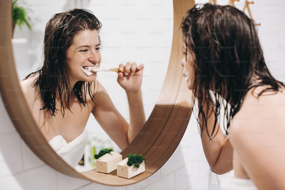 Tooth care concept. Young happy woman in white towel brushing teeth and looking at round mirror in stylish bathroom. Slim sexy woman daily routine after shower.