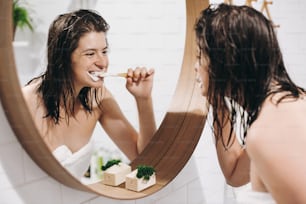 Concepto de cuidado dental. Joven mujer feliz con toalla blanca cepillándose los dientes y mirando el espejo redondo en el baño elegante. Rutina diaria de la mujer delgada y sexy después de la ducha.