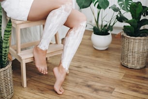 Young woman in white towel applying shaving cream on her legs in home bathroom with green plants. Skin care and wellness. Hair Removal concept, depilation cream.