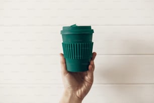 Verbot von Einwegplastik. Hand hält stilvolle wiederverwendbare Öko-Kaffeetasse auf weißem Holzhintergrund. Green Cup aus natürlicher Bambusfaser, Zero-Waste-Konzept. Treffen Sie eine Wahl. Kaffee zum Mitnehmen