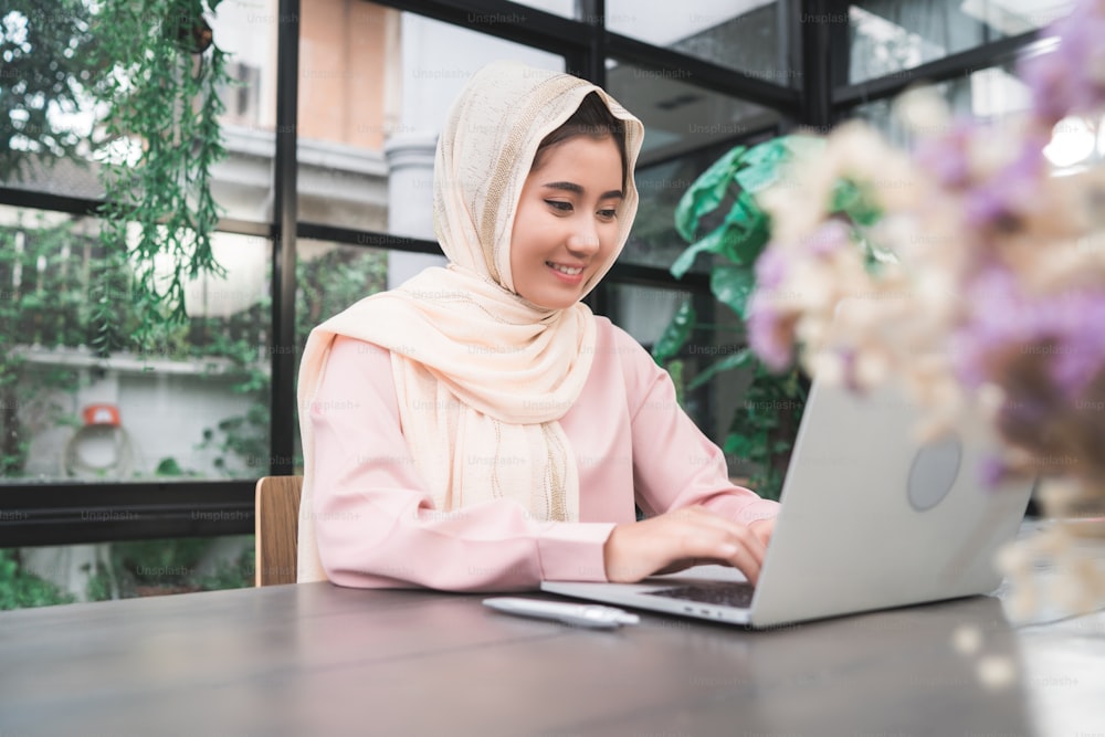 Schöne junge lächelnde asiatische muslimische Frau, die am Laptop arbeitet, sitzt im Wohnzimmer zu Hause. Asiatische Geschäftsfrau arbeitet an Dokumentenfinanzierung und Taschenrechner in ihrem Heimbüro. Die Zeit zu Hause genießen.