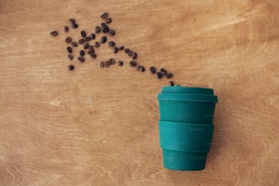Zero-Waste-Konzept, flache Verlegung. Stilvolle wiederverwendbare Öko-Kaffeetasse auf Holzhintergrund mit gerösteten Kaffeebohnen. Verbot von Einwegplastik. Nachhaltiger Lebensstil. Natürlicher Bambusbecher
