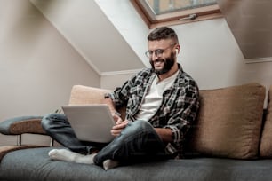 Tan cómodo. Hombre alegre y feliz sentado en el sofá con su computadora portátil mientras trabaja en él