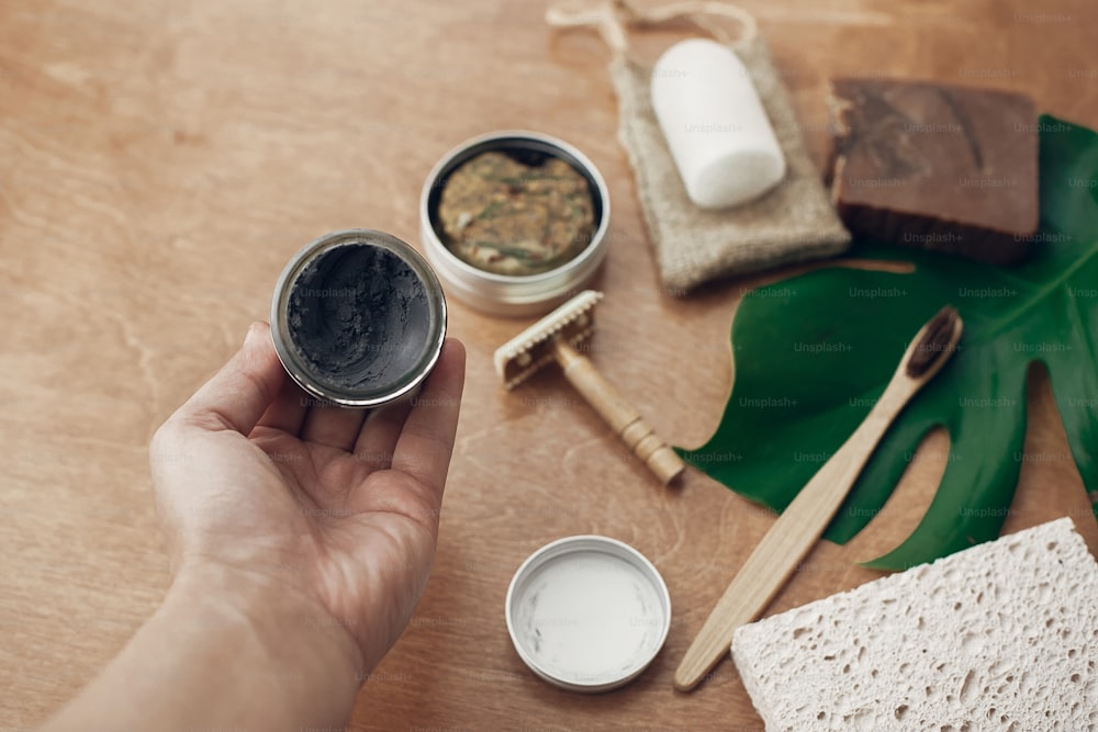 Zero waste. Hand holding natural eco charcoal toothpaste in glass jar on background of bamboo toothbrush, soap, solid shampoo, metal razor, deodorant on wood with green monstera leaf.