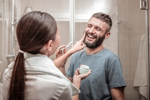 Caring girlfriend. Nice pleasant woman standing in front of her boyfriend while putting cream on his face