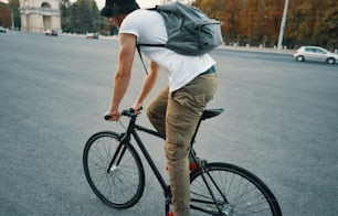 Side back view young modern man, student with backpack, cycling on a classic bike on the city road. Copy space. Active and ecological lifestyle concept