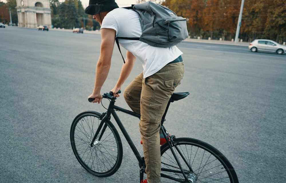 Side back view young modern man, student with backpack, cycling on a classic bike on the city road. Copy space. Active and ecological lifestyle concept