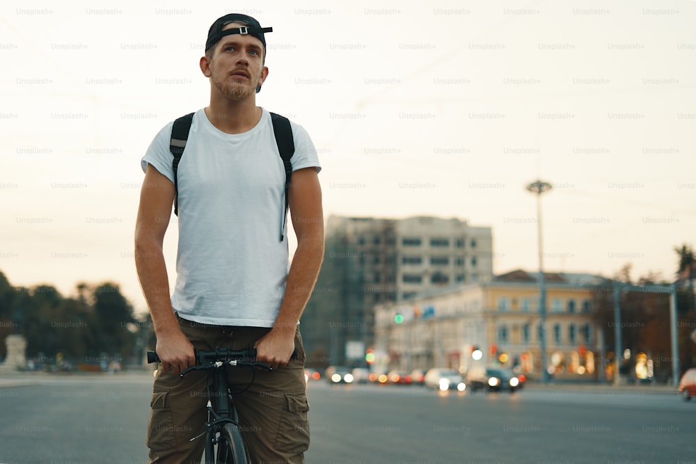 Retrato de un joven montando en bicicleta en la carretera de la ciudad, calle con la ciudad lejos en el fondo. Hombre en bicicleta negra con camisa blanca, gorra, mochila pedaleando hacia el destino. Concepto de estilo de vida saludable