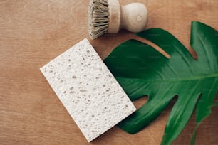 Zero waste concept, flat lay. Reusable natural cellulose sponge and eco wooden brush on wooden background with green monstera leaf. Ban single use plastic. Sustainable lifestyle.