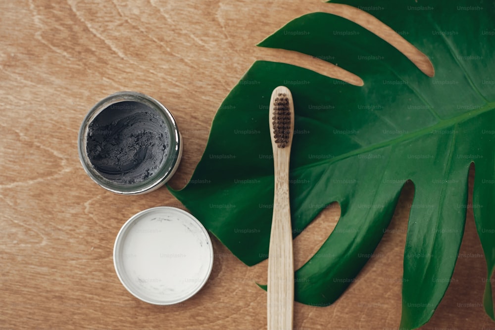 Natural toothpaste activated charcoal and bamboo toothbrush on wooden background with green monstera leaf. Plastic free beauty essentials, teeth care. Zero waste concept. Sustainable lifestyle
