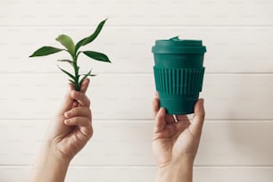 Manos sosteniendo una elegante taza de café ecológica reutilizable y hojas de bambú verde sobre fondo de madera blanca. Residuo cero. Vaso verde de fibra de bambú natural. Prohibir el plástico de un solo uso. Toma una decisión