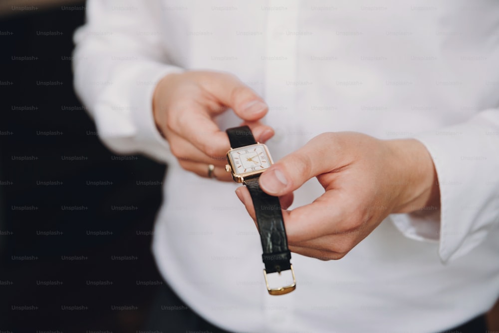 Businessman holding luxury watch. Stylish groom putting on white shirt and watch, getting ready in the morning near window in soft light, before wedding ceremony.  Time is money