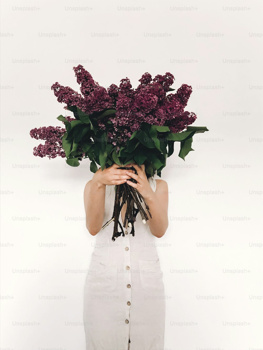 Girl in rustic linen dress holding purple lilac flowers in front of face on white background. Hello spring. Happy Mothers day. Lilac bouquet in woman hands. International womens day