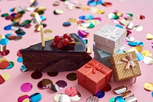 Una fetta di torta al cioccolato con candelina e piccole scatole regalo per il tuo compleanno. Sfondo rosa con coriandoli