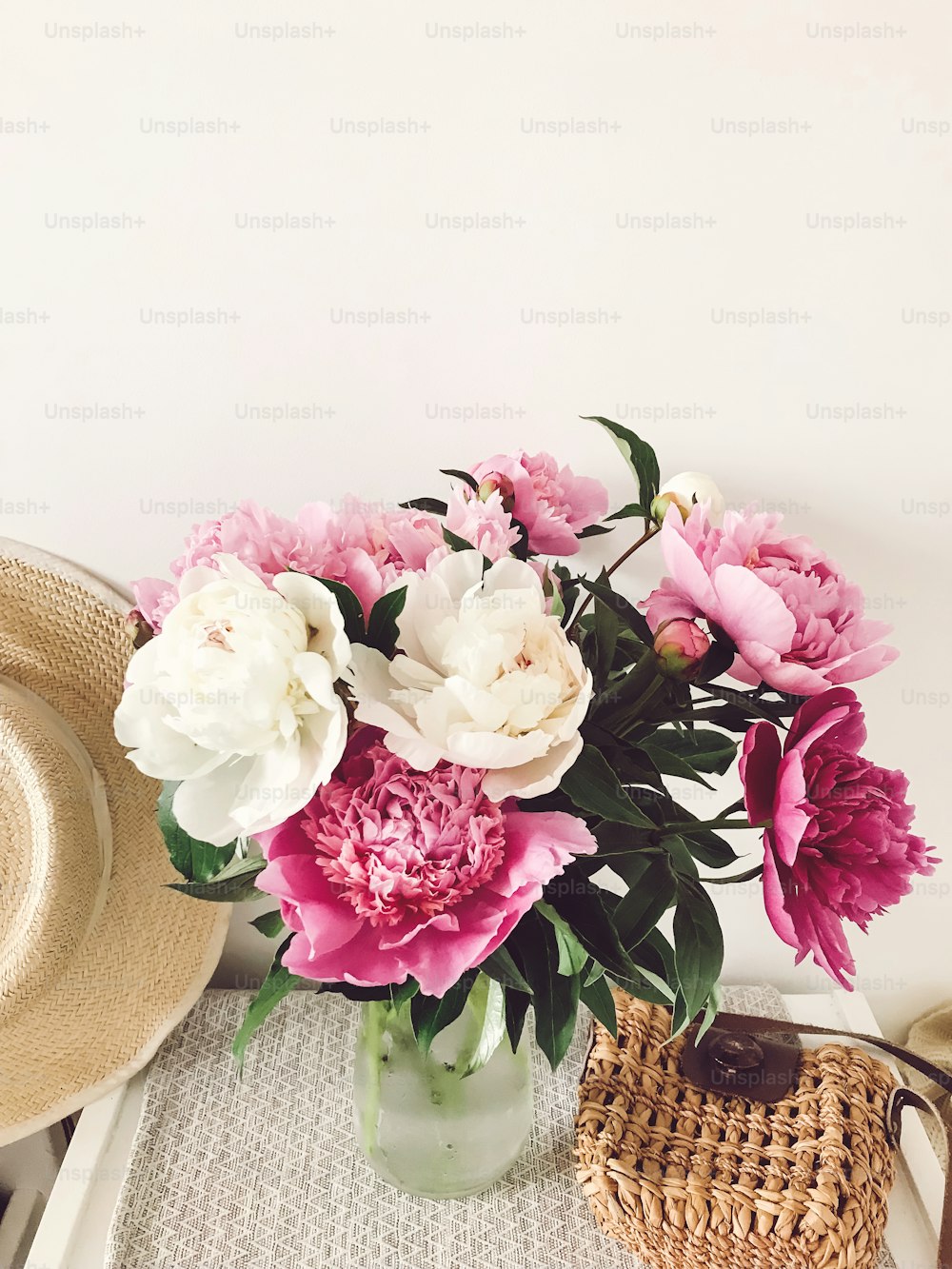 Stylish pink and white peonies in vase and trendy straw bag and hat on stylish white nightstand near bed. Hello spring. Happy Mothers day. Girly image