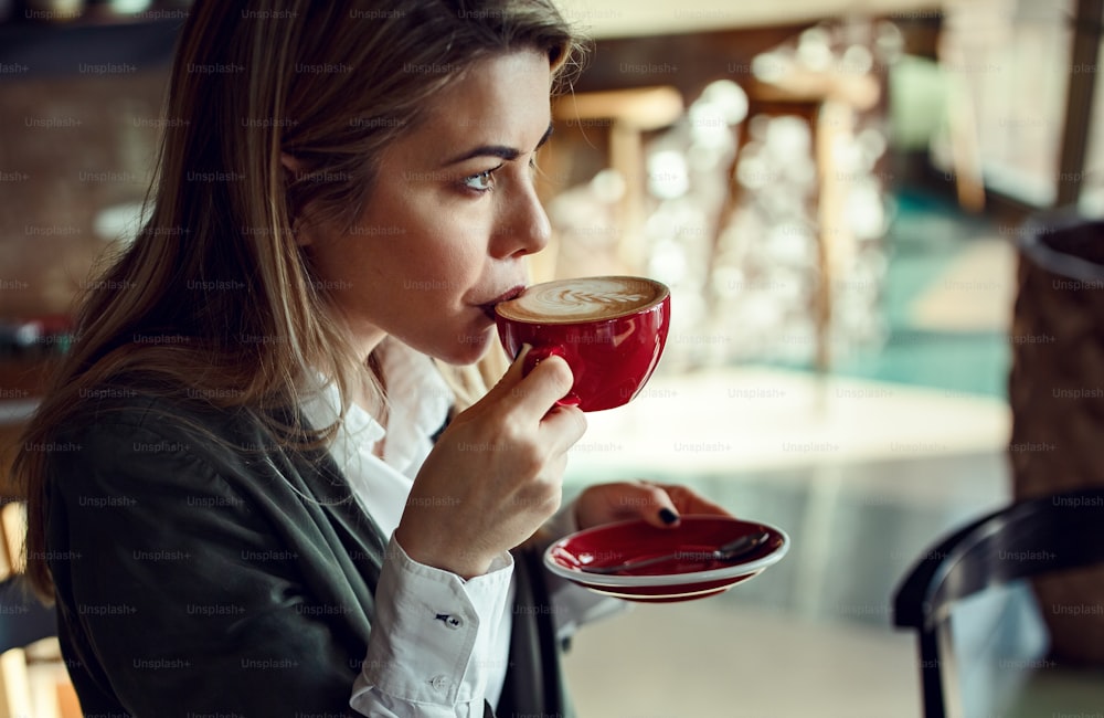カフェで一人で座りながらラテコーヒーの味を楽しむ若い思慮深いビジネスウーマン。