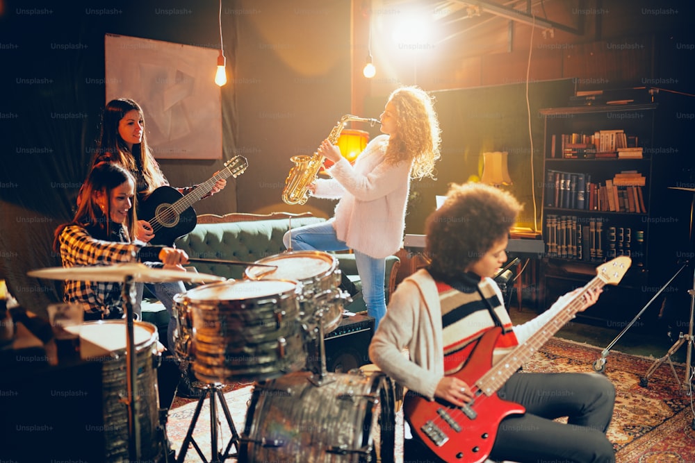 Girls playing jazz music. In foreground one woman playing bass guitar and in background other three playing acoustic guitar, saxophone and drums. Home studio interior.
