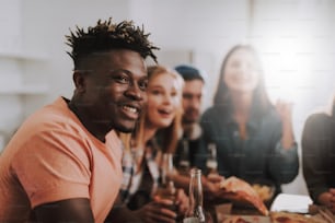 Portrait of joyful young man with dreadlocks looking away and smiling while sitting at the table. Two girls and gentleman on blurred background