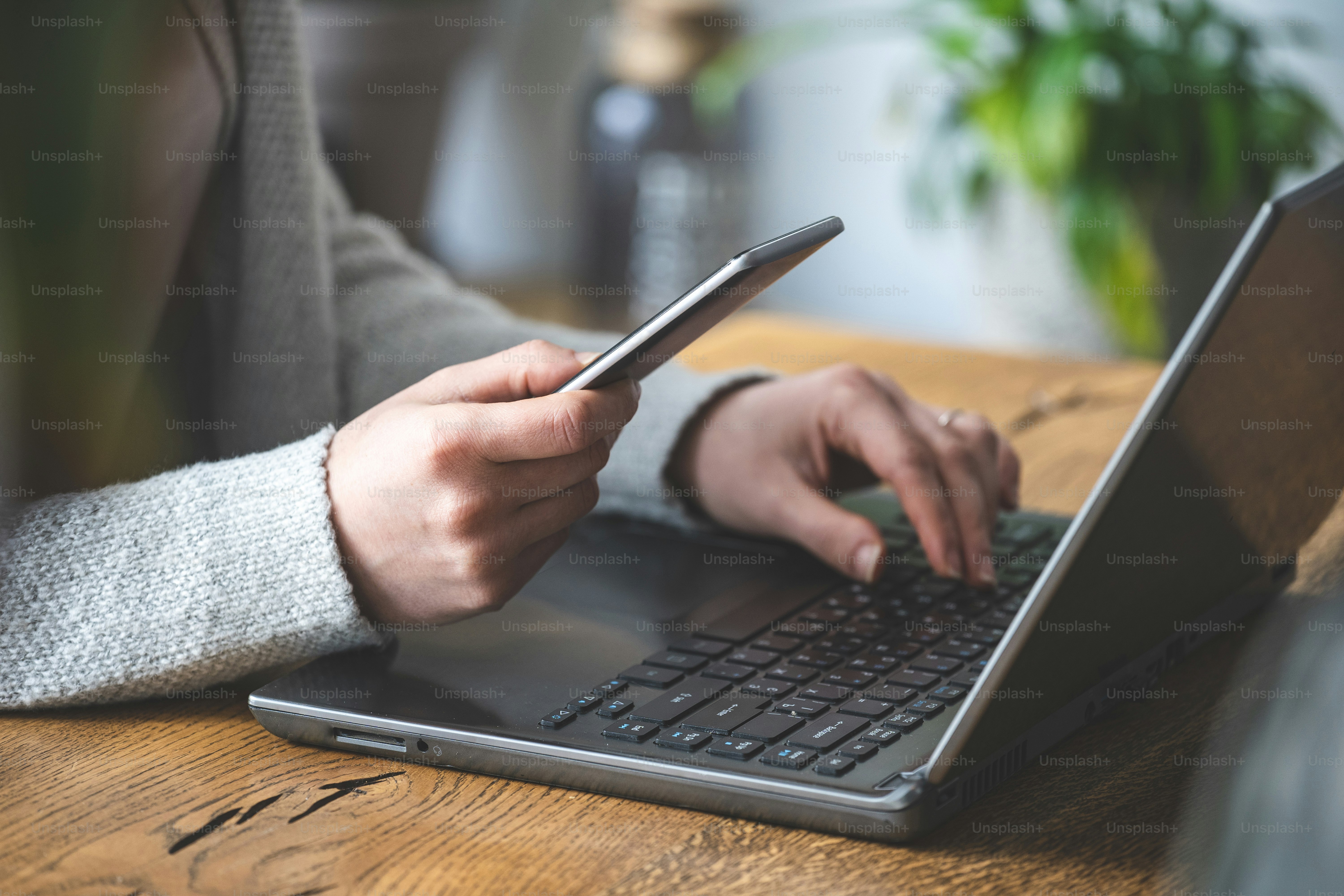 Nahaufnahme und beschnittenes Foto einer geschäftigen, nicht erkennbaren Dame. Sie sitzt im Loft-Innenraum und benutzt einen Laptop und hält tragbare Telefongeräte in ihren Händen.