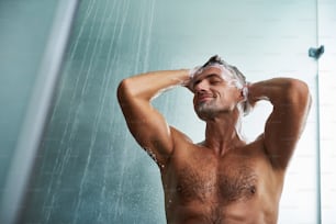 Ritratto ad angolo basso di attraente gentiluomo nudo che fa la doccia a casa. Chiude gli occhi con piacere e sorride