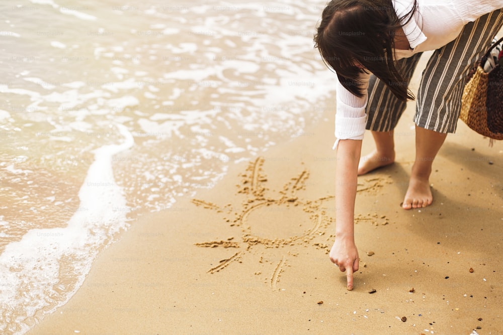 Young woman writing sun on sand beach near sea and waves. Hipster girl relaxing on sandy beach, walking and having fun. Summer vacation concept