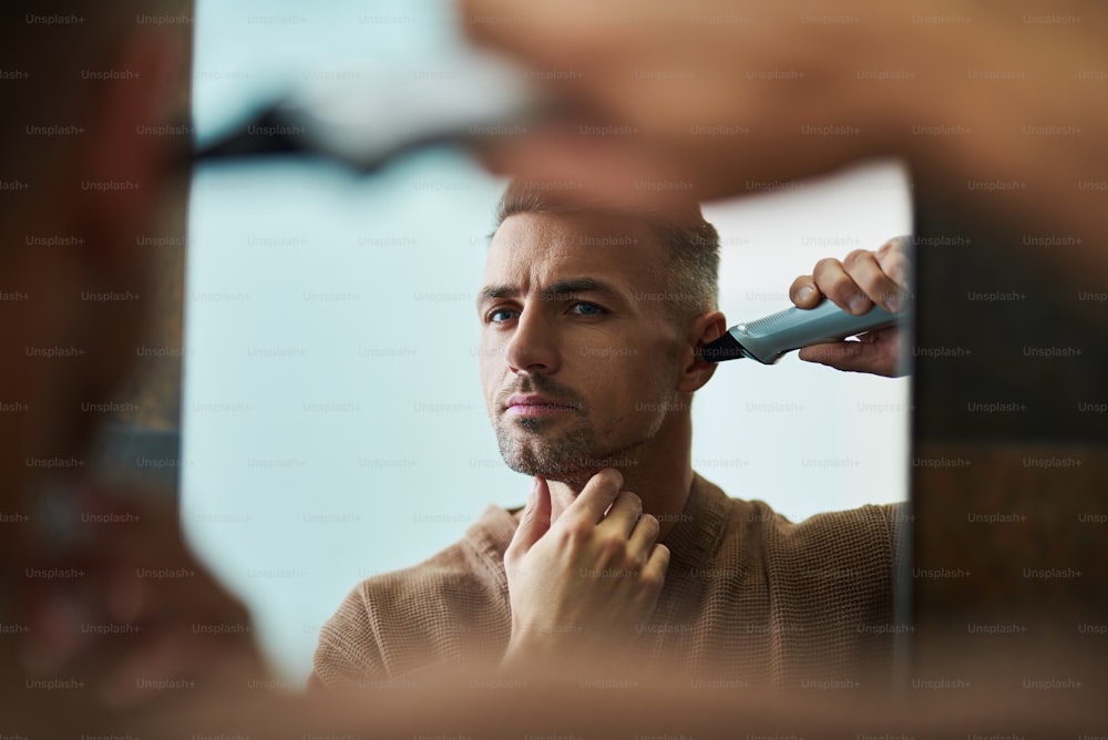 Portrait of good-looking gentleman in bathrobe using electric trimmer while looking in the mirror with serious expression