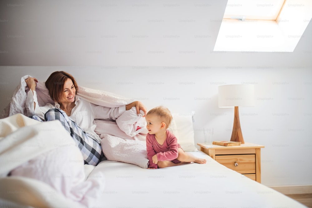 Uma jovem mãe feliz com a filhinha sentada dentro de casa na cama de manhã, brincando.