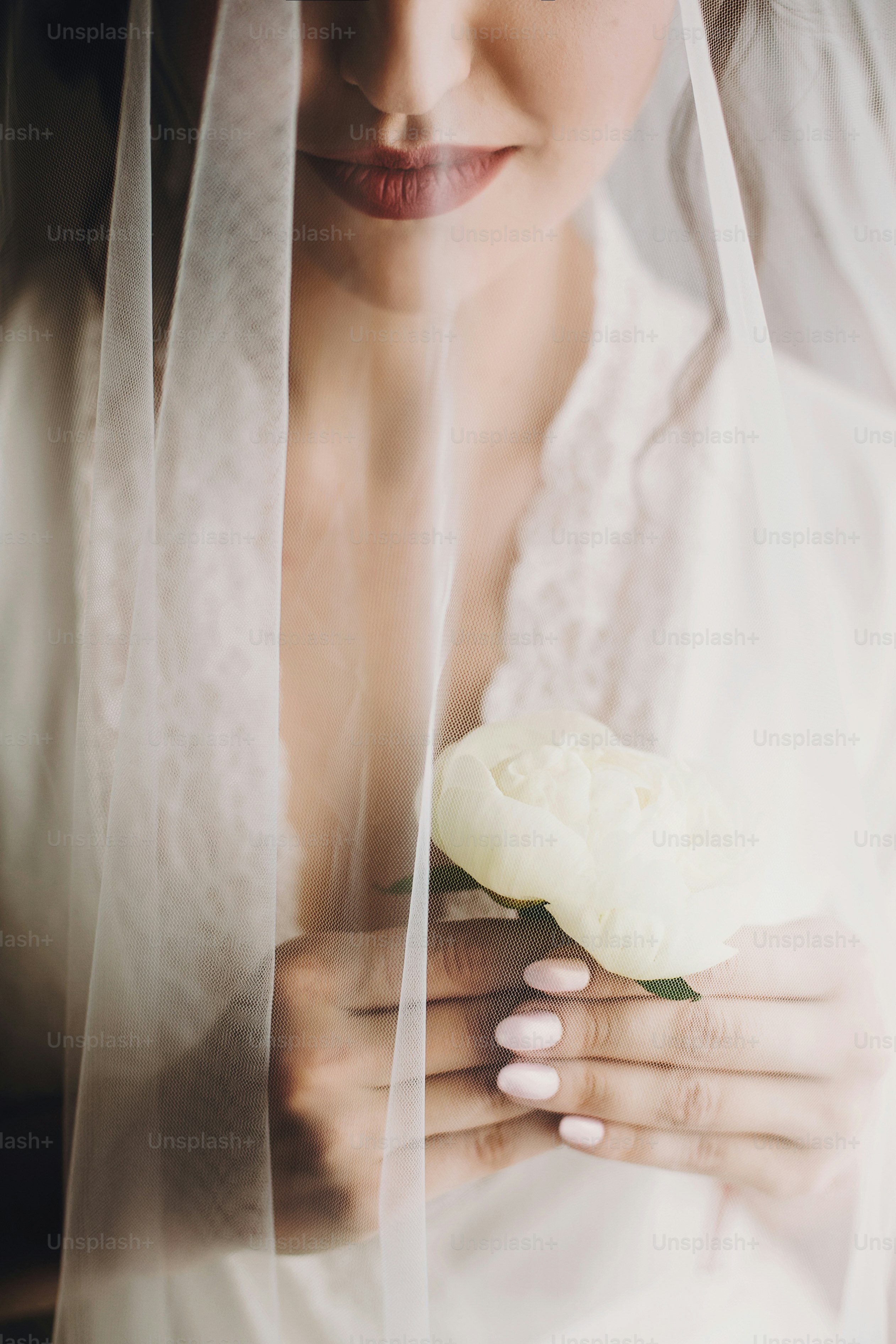 Beautiful stylish brunette bride posing in silk robe under veil in the morning. Sensual portrait of happy woman model with peony flower in hands getting ready for wedding day