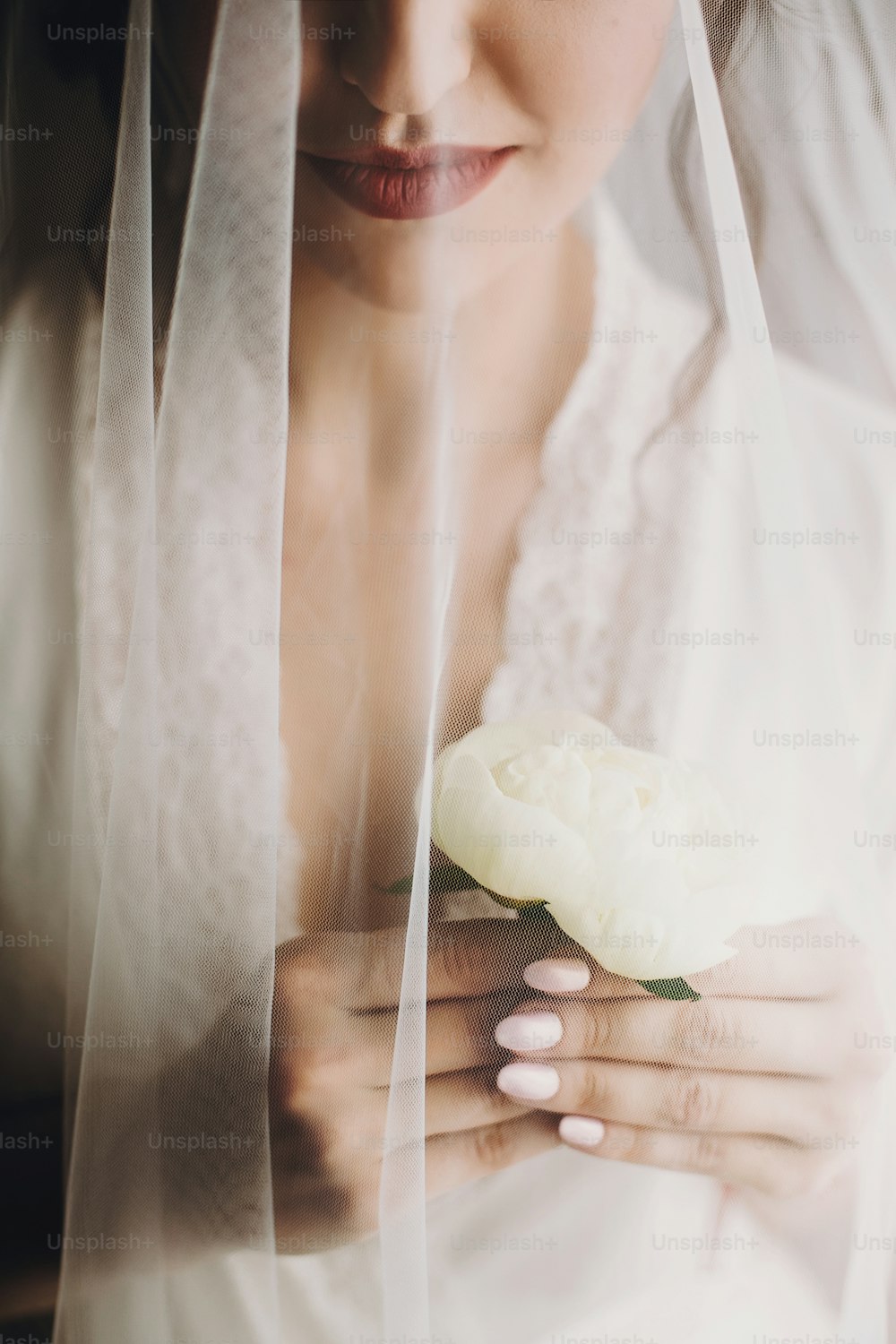 美しいスタイリッシュなブルネットの花嫁は、朝にベールの下にシルクローブでポーズをとっています。結婚式の日の準備をしている手に牡丹の花を持つ幸せな女性モデルの官能的な肖像画