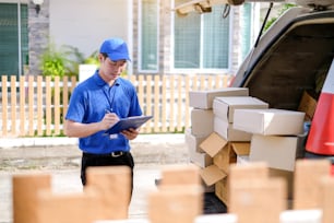 Jeune livreur en uniforme bleu vérifiant les boîtes de produits à envoyer aux clients dans des véhicules de transport.