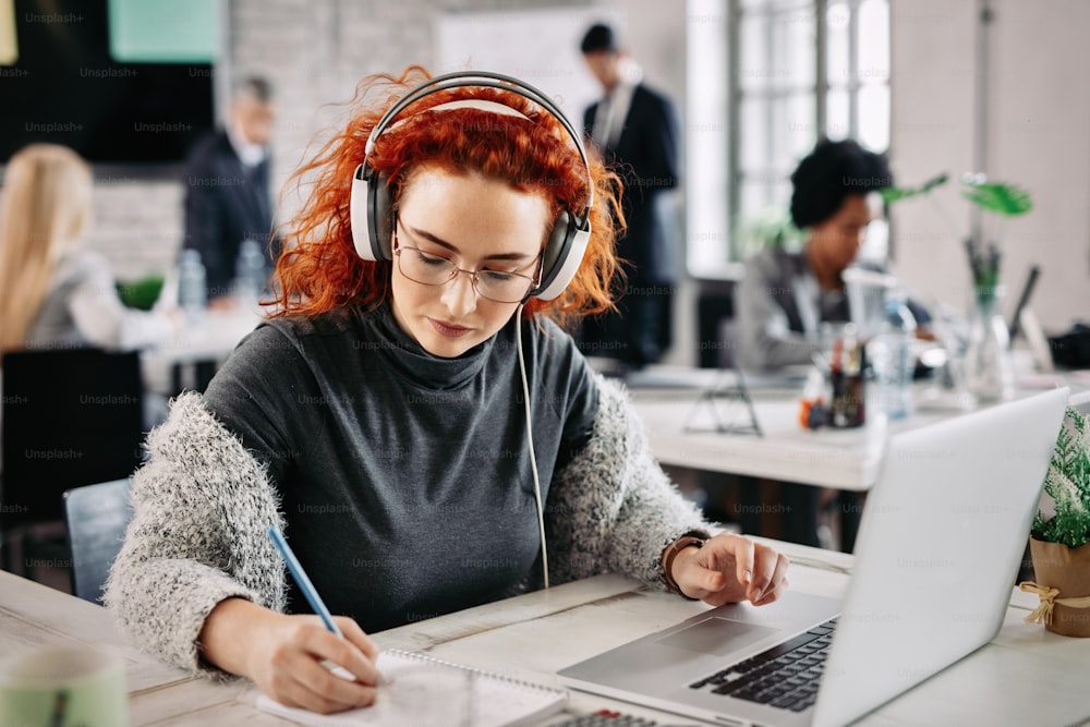 Empresária ruiva usando laptop e escrevendo anotações em seu bloco de notas enquanto ouve música em fones de ouvido no trabalho.