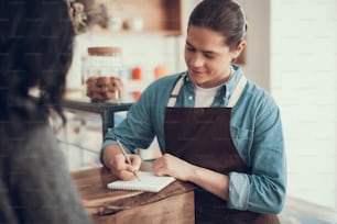 Attentive experienced waiter putting on elbow on the bar counter and slightly smiling while writing the order to his notebook