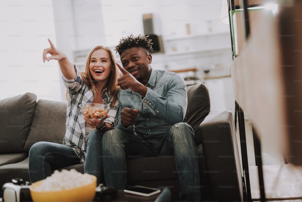 Having fun at home. Portrait of charming young lady holding bowl with snacks while sitting on couch with handsome afro american guy