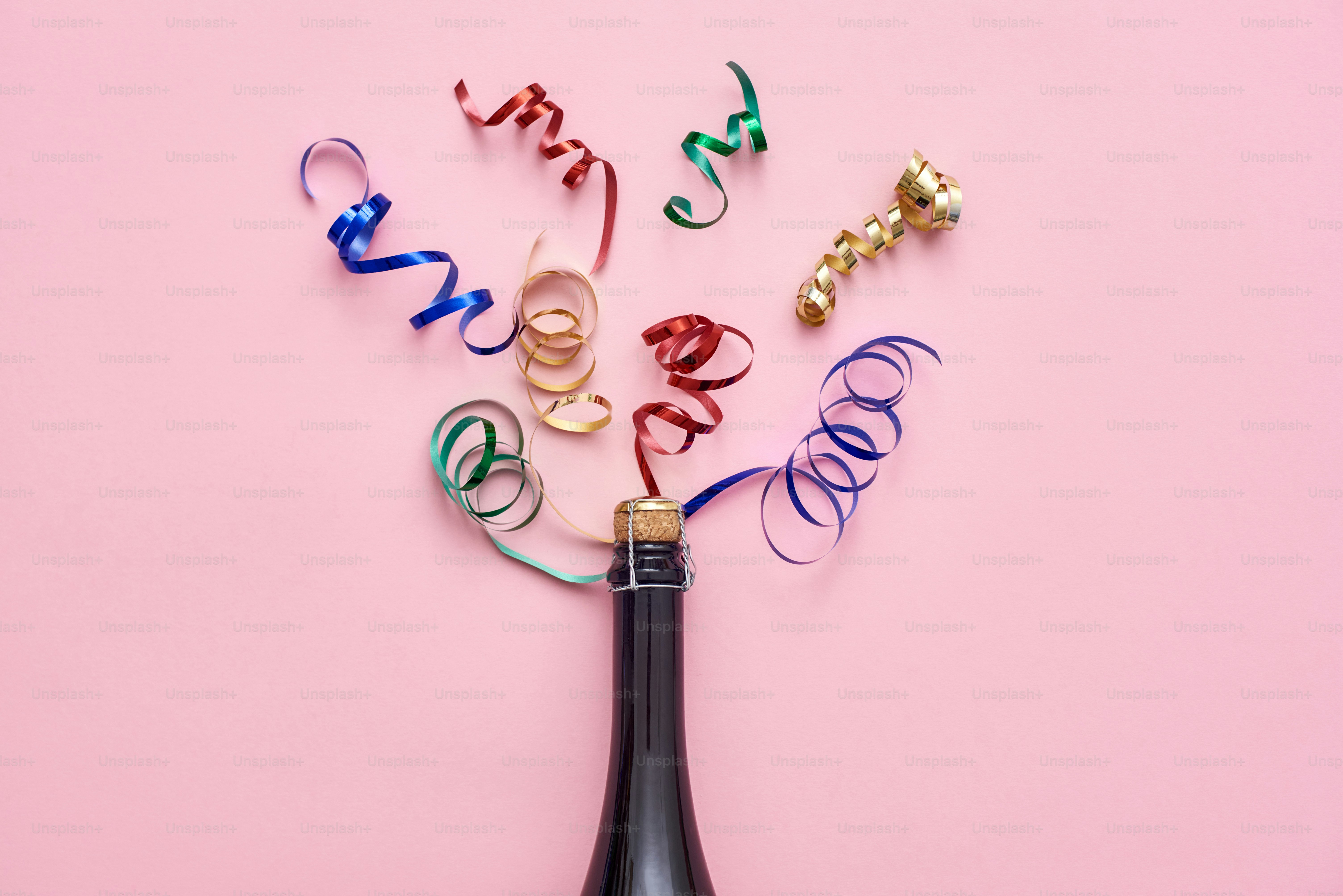 Party up. A bottle of champagne with shiny ribbons on pink background. Top view