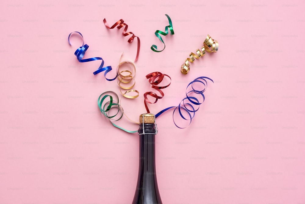 Party up. A bottle of champagne with shiny ribbons on pink background. Top view