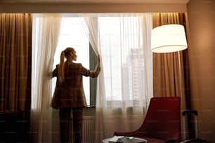 Confortevole camera d'albergo. La donna d'affari nella stanza d'albergo buia apre le tende sulla finestra alla luce del mattino