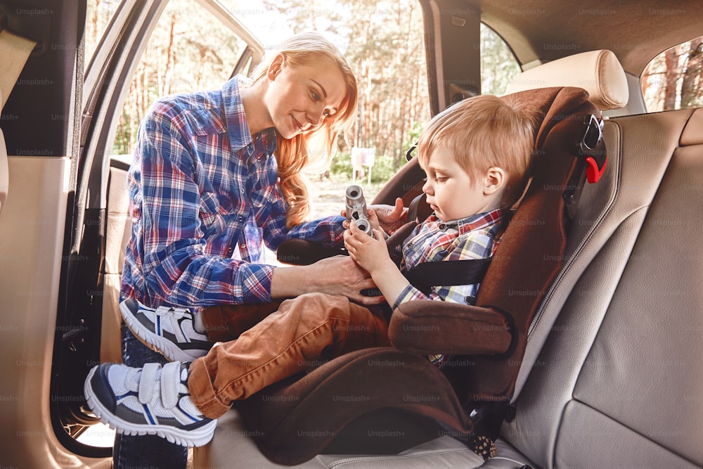 La mère attache le siège d’auto de l’enfant, tandis que l’enfant joue avec des jumelles. Vue latérale