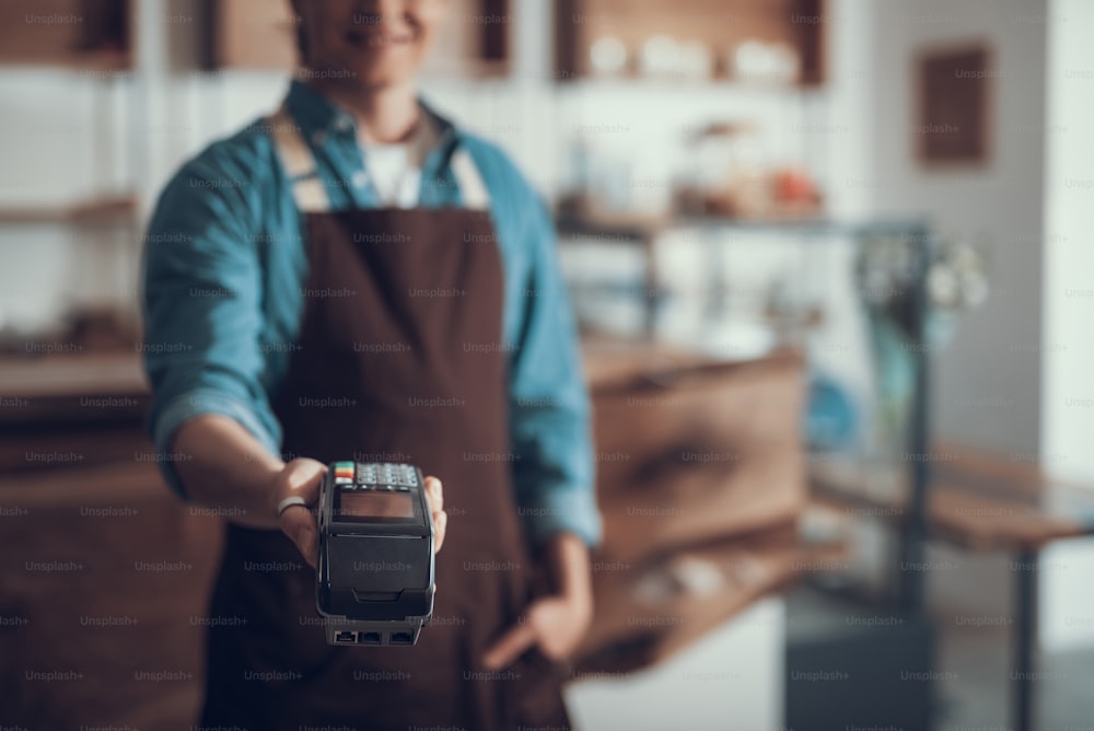 Jeune serveur professionnel debout dans le café et mettant sa main en avant tout en tenant la machine de paiement par carte
