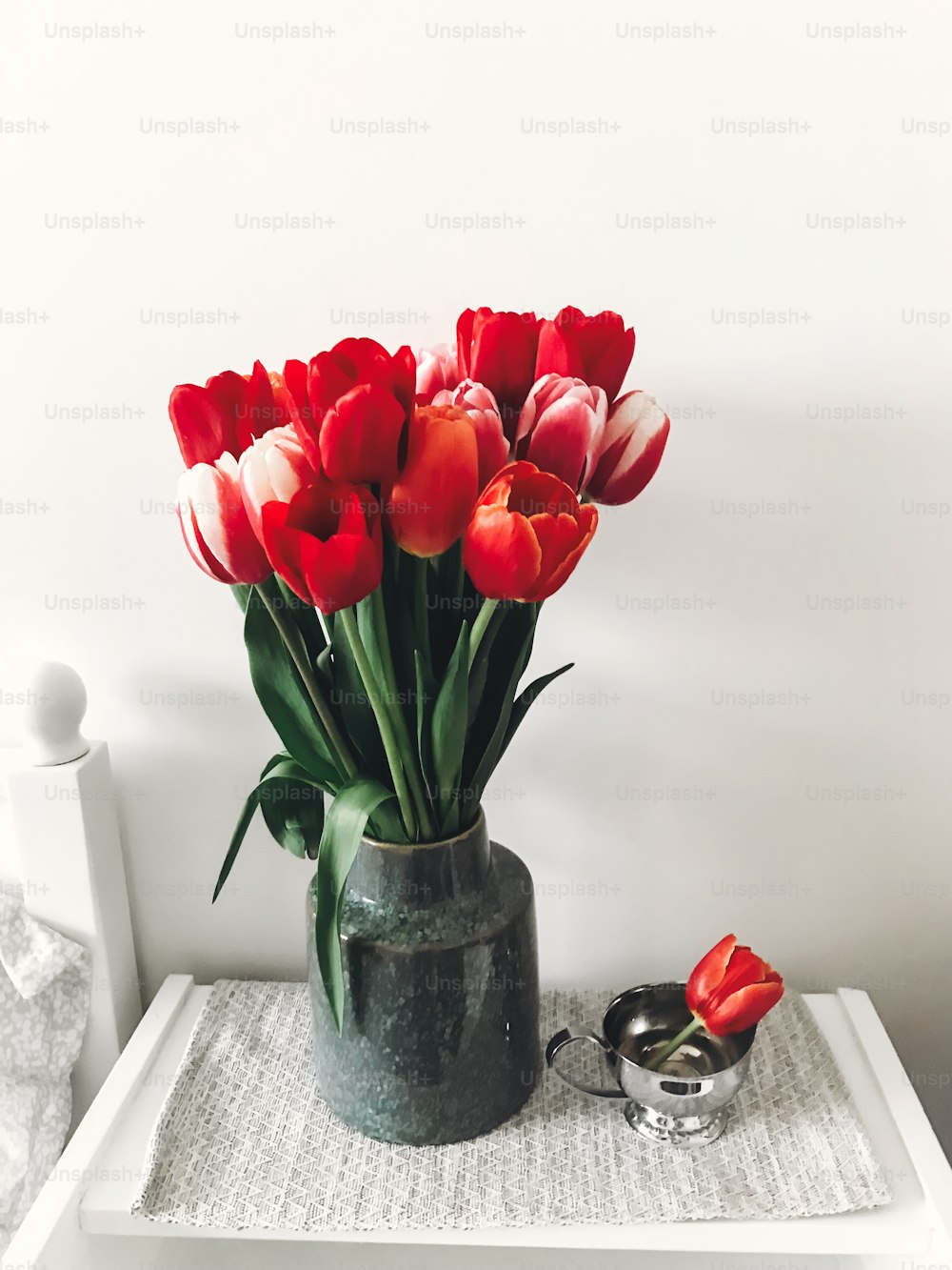 Beautiful red and pink tulips in vase on white wooden nightstand near stylish white bed, copy space. Hello spring concept. Happy mothers day