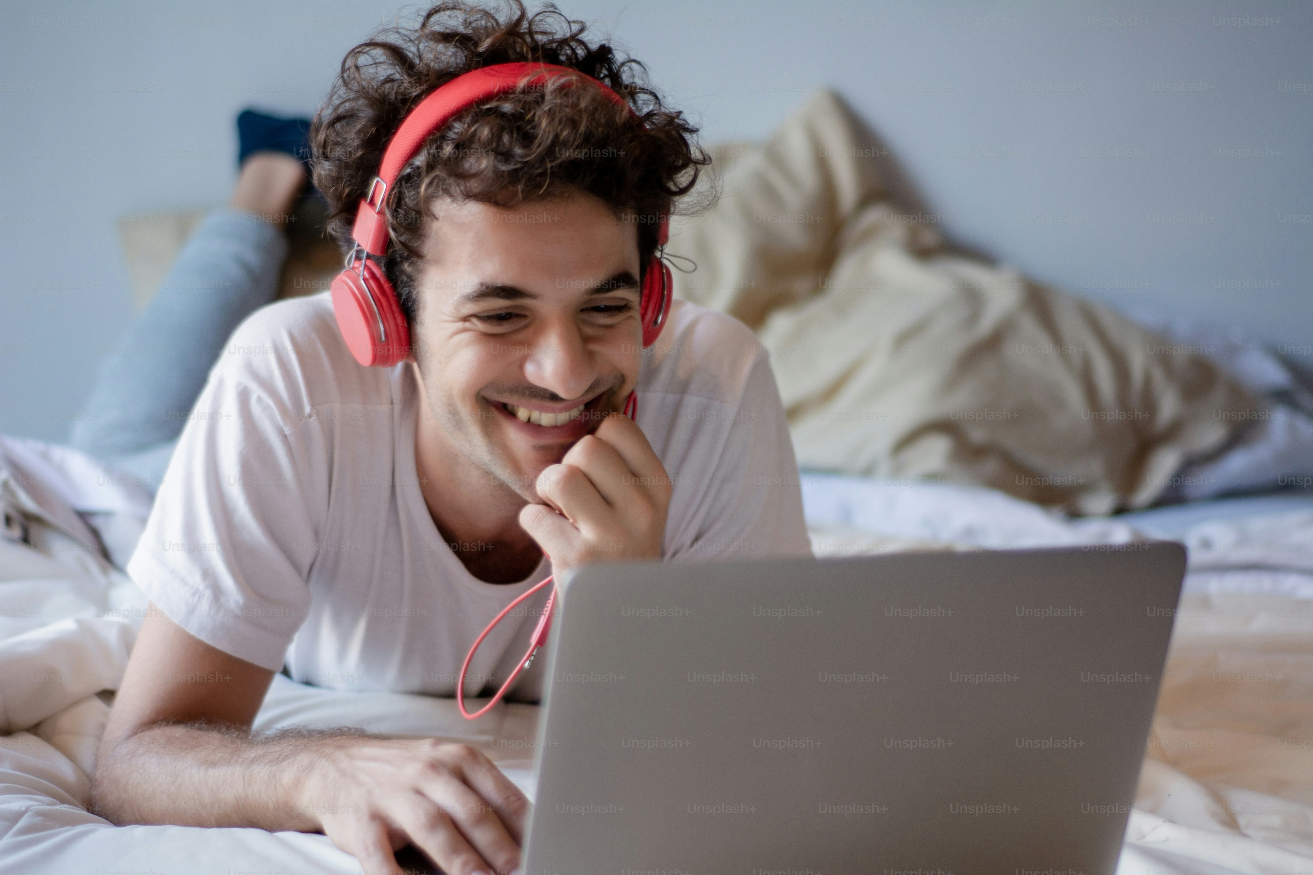 Pemuda bahagia mendengarkan musik dengan headphone dan menggunakan laptop di tempat tidurnya. Di dalam ruangan