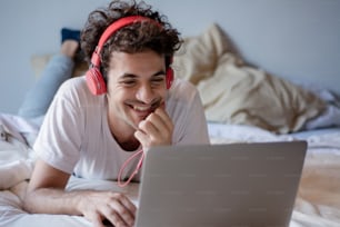 Happy young man listening music with headphones and using laptop on his bed. Indoors