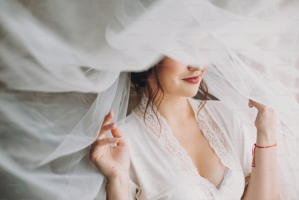 Schöne stilvolle brünette Braut posiert morgens in Seidenrobe unter Schleier. Glückliches Frauenmodel mit perfektem Make-up und Frisur, bereitet sich auf den Hochzeitstag vor